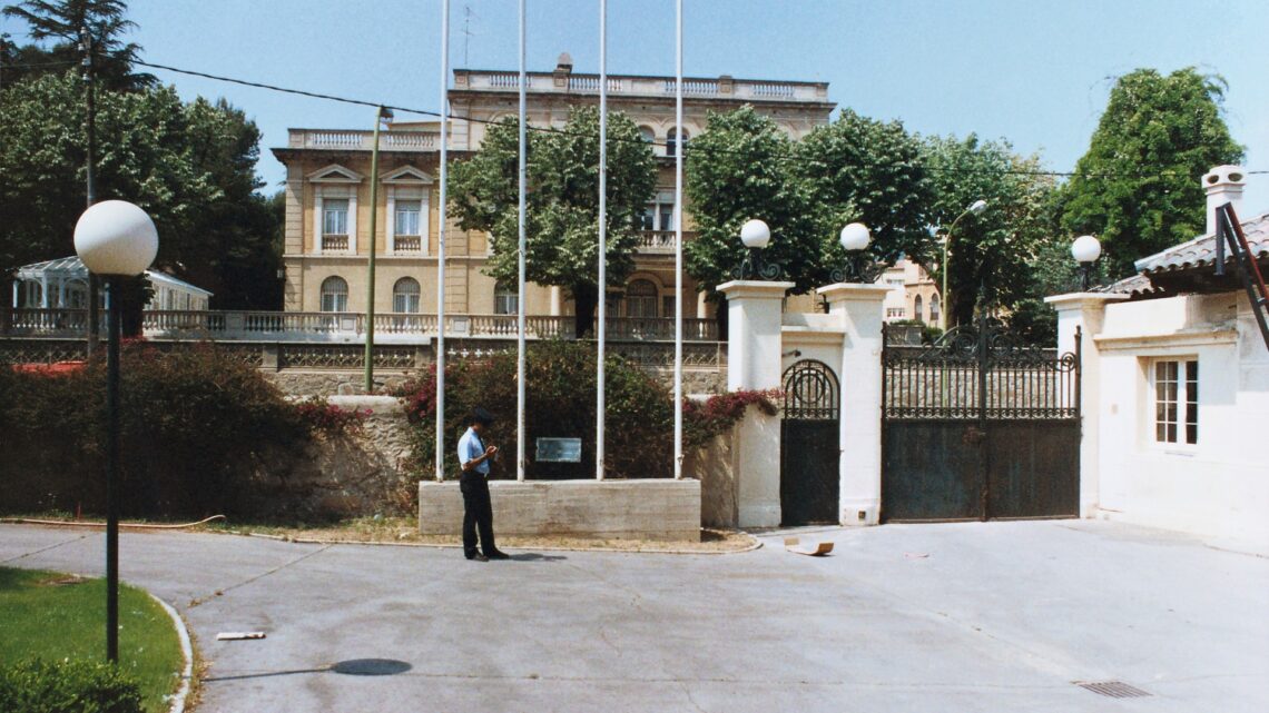 A formal consulate building with gates, flagpoles, and a uniformed guard
