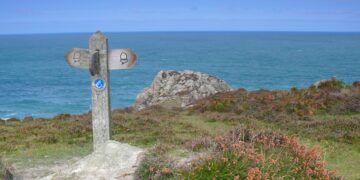 A wooden waymarker sign on the Pembrokeshire Coast Path overlooking the sea and coastal rock outcrops