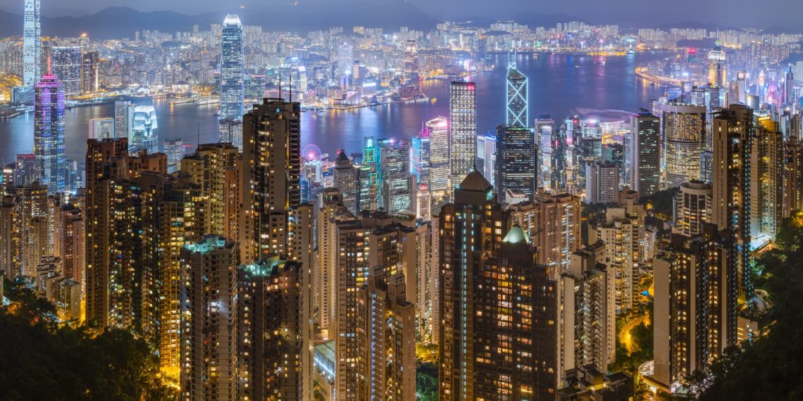Dense residential apartment towers in the foreground with Victoria Harbour and Hong Kong's skyline behind, seen at night from Victoria Peak