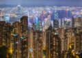 Dense residential apartment towers in the foreground with Victoria Harbour and Hong Kong's skyline behind, seen at night from Victoria Peak