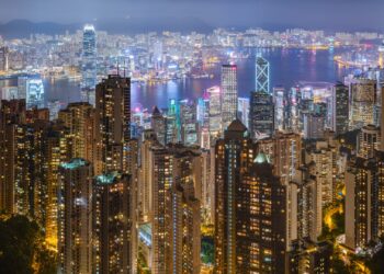 Dense residential apartment towers in the foreground with Victoria Harbour and Hong Kong's skyline behind, seen at night from Victoria Peak