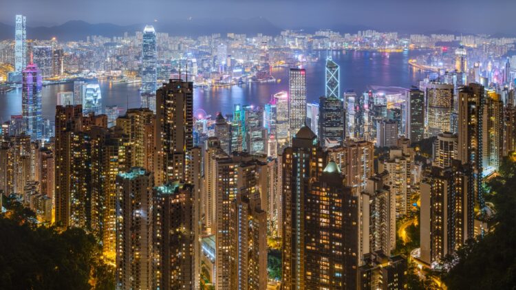 Dense residential apartment towers in the foreground with Victoria Harbour and Hong Kong's skyline behind, seen at night from Victoria Peak