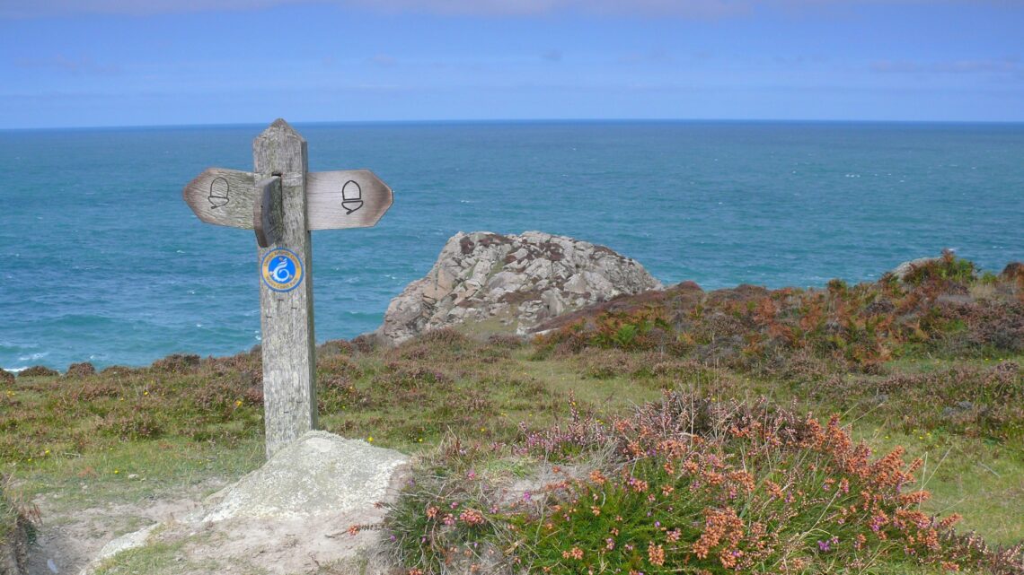 A wooden waymarker sign on the Pembrokeshire Coast Path overlooking the sea and coastal rock outcrops